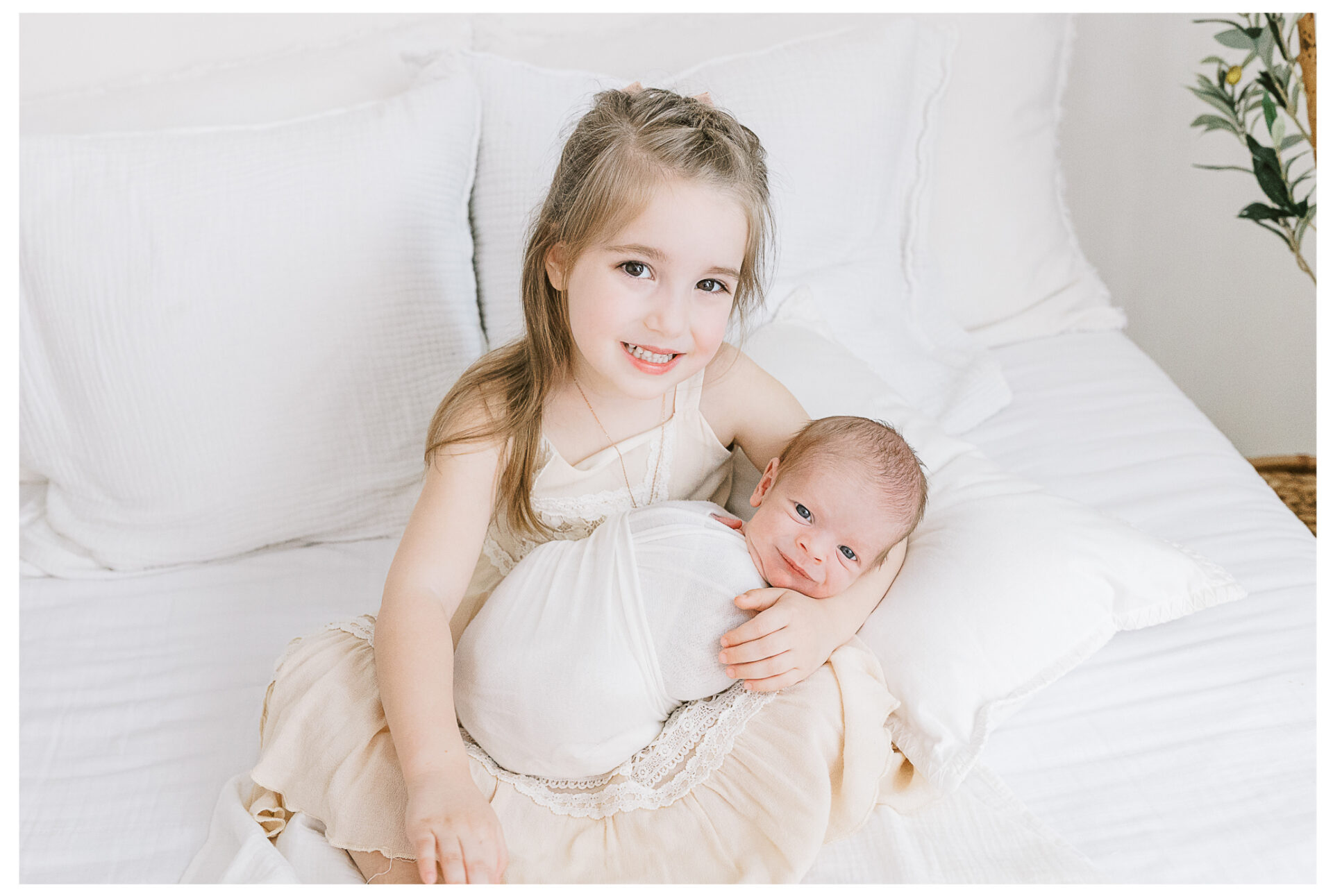A young girl in a cream dress sits on a white bed, smiling at the camera and gently holding her newborn brother wrapped in a white blanket on her lap. Bright and soft natural light fills the room.