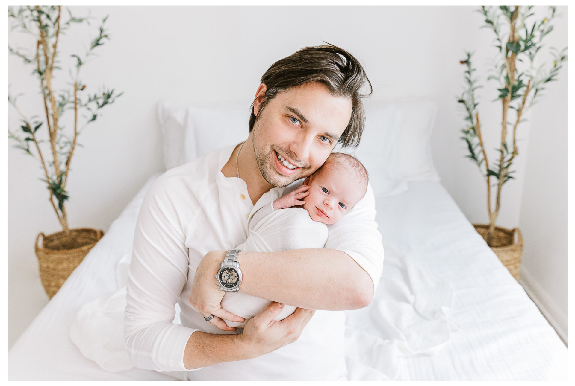 A father in a white shirt sits on a bed, smiling and holding his newborn baby wrapped in a white blanket. Two potted plants are visible in the background. The room is bright and airy.