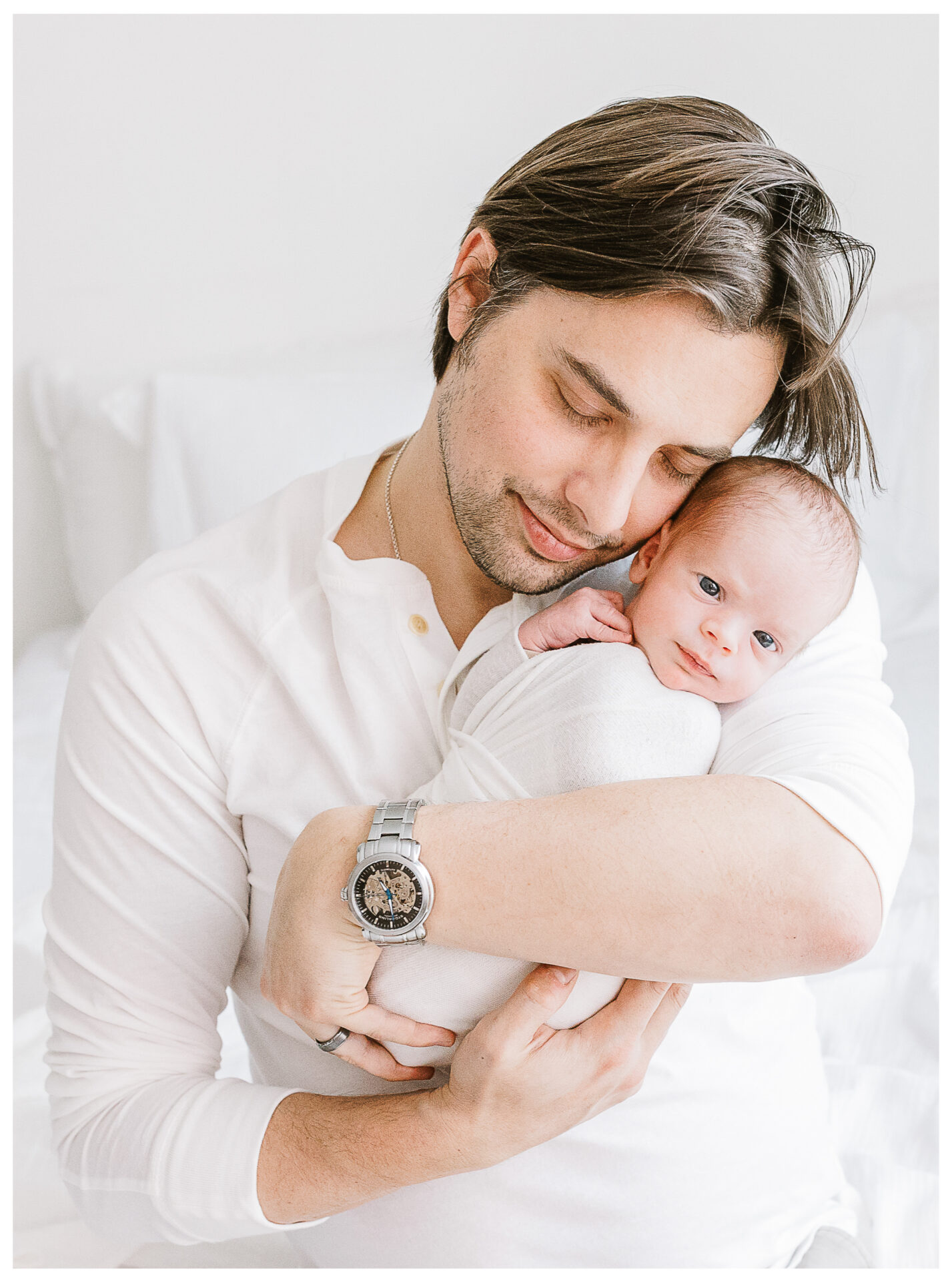 A father in a white shirt lovingly cradles his swaddled newborn baby in his arms, both resting on a white bed, with the man looking down and smiling gently while the baby gazes at the camera.