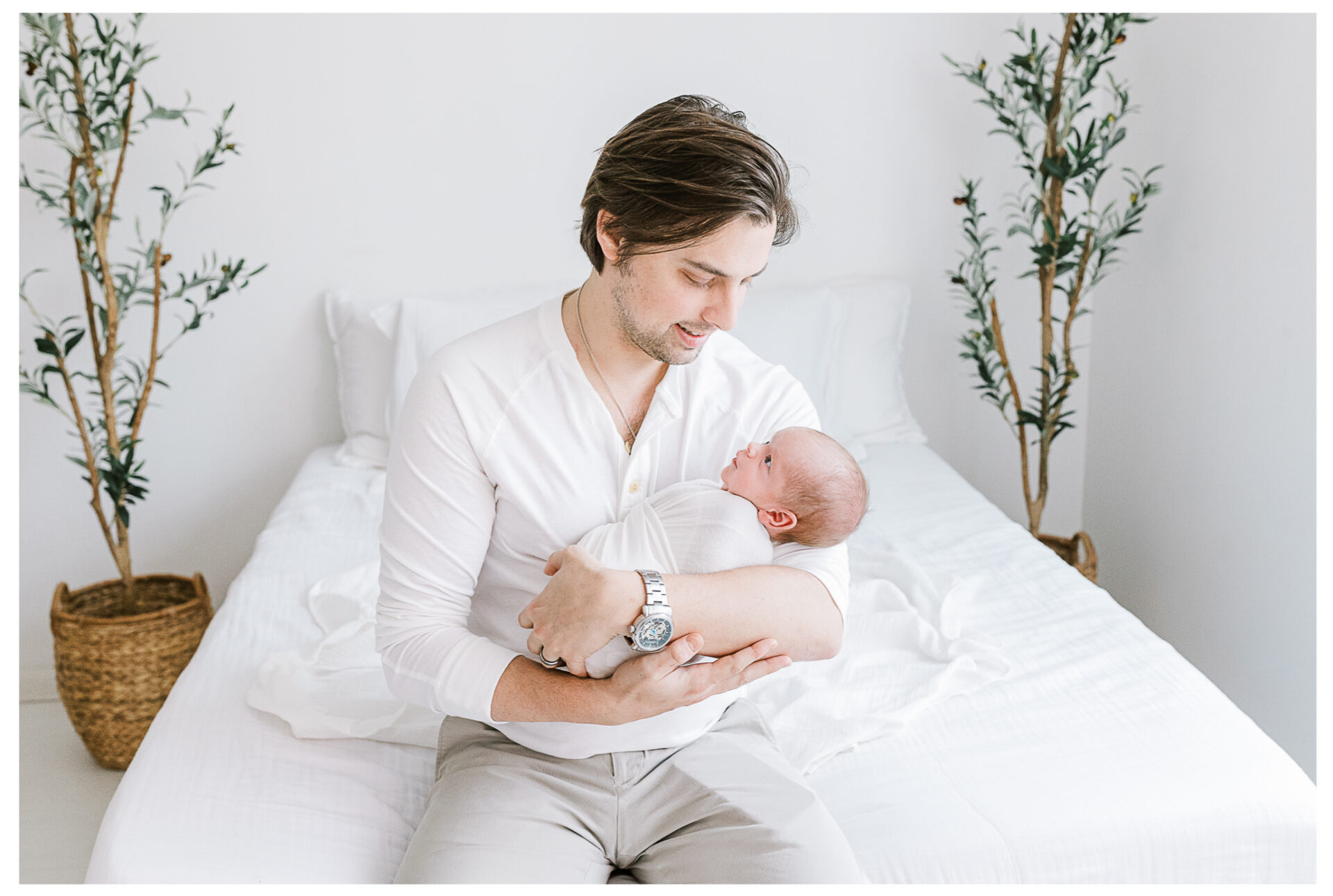 A father in a white shirt sits on a white bed, gently cradling his swaddled baby boy in his arms. Two potted plants are placed on either side of the bed, adding greenery to the bright, minimal room.