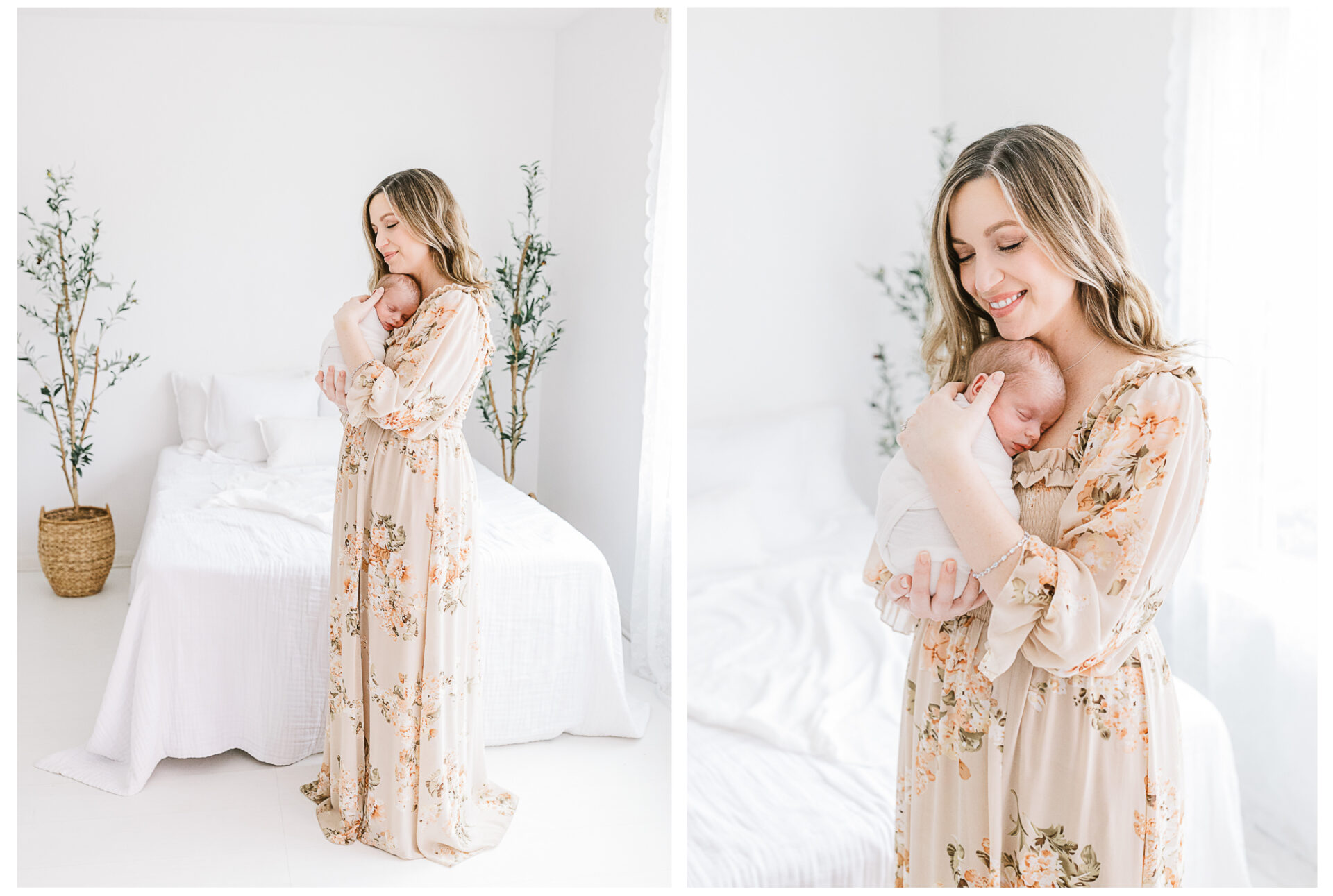 A mother in a light floral dress gently holds her sleeping newborn wrapped in a white blanket, standing beside a white bed in a bright, airy room with minimal decor and green plants.