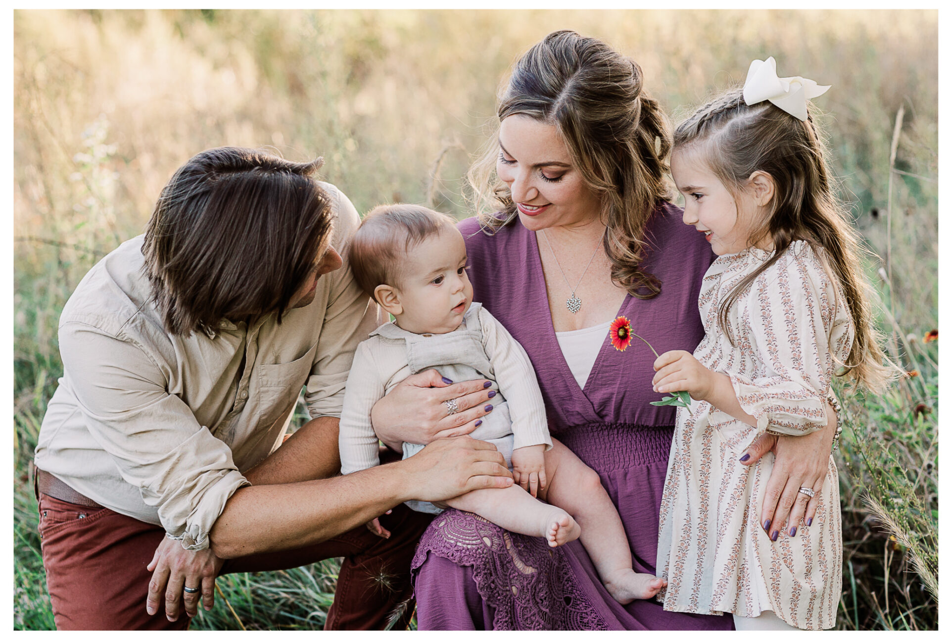 A family of four sits together outdoors in a grassy field. The mother in a purple dress holds her baby boy, the father snuggles close and their young daughter with a white bow in her hair leans in, all smiling and sharing a tender moment.