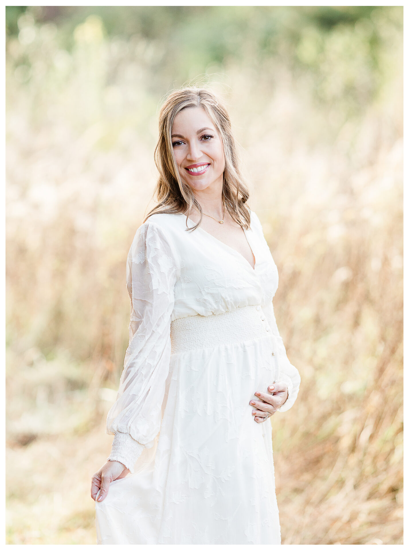 A smiling woman in a long, white, flowy dress stands outdoors in a sunlit, grassy field, gently holding her dress with one hand and resting her other hand on her belly. The background is softly blurred.