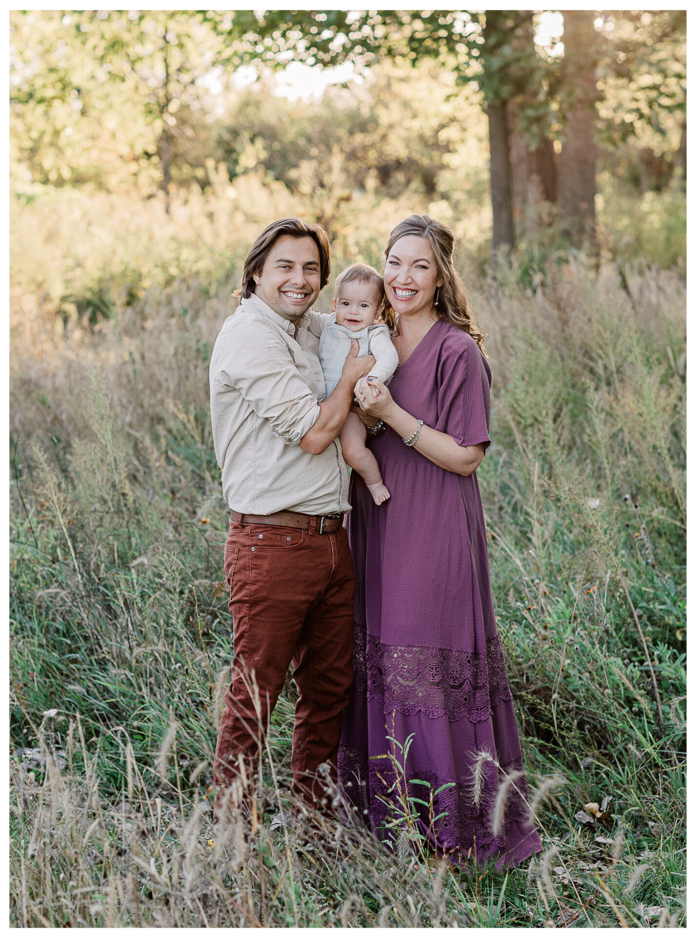 A smiling husband and wife stand in a grassy field holding their baby boy between them. The man wears a tan shirt and rust-colored pants; the woman, a purple dress. Tall grass and fall trees brighten the outdoor background.
