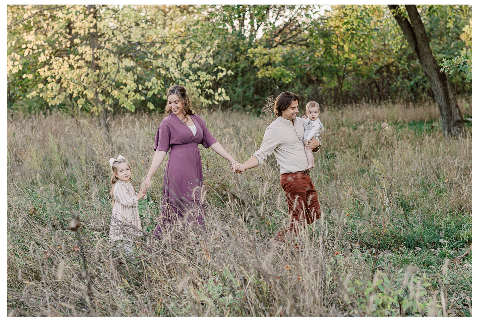 A family of four walks through tall grass in a fall field. The mother in a purple dress holds her daughter's hand, while the father, wearing a beige shirt, holds his baby boy and walks beside them. Trees with autumn leaves are in the background.