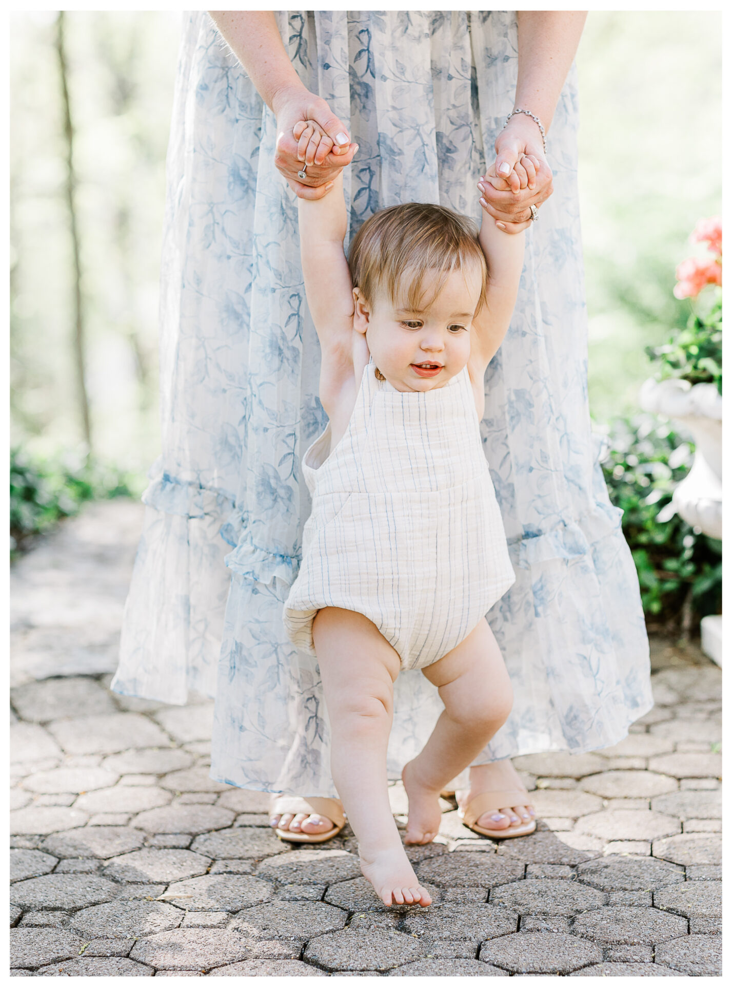 A baby in a plaid romper takes steps while holding an adult’s hands. Part of the Dayton Ohio baby portrait collection, this sunny outdoor moment captures them on a stone path with greenery in the background.