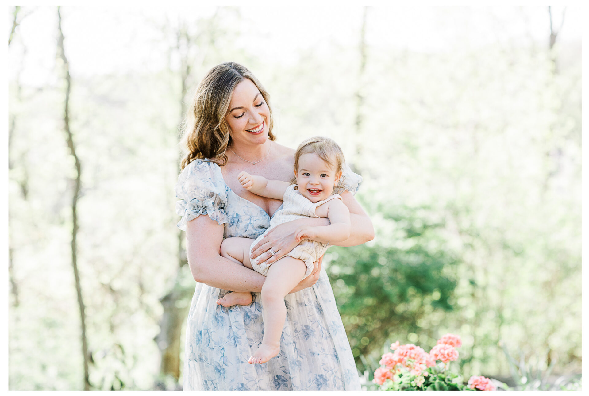 A mother in a light floral dress smiles while holding her laughing baby outdoors. The background is bright and blurred with green foliage and pink flowers visible.
