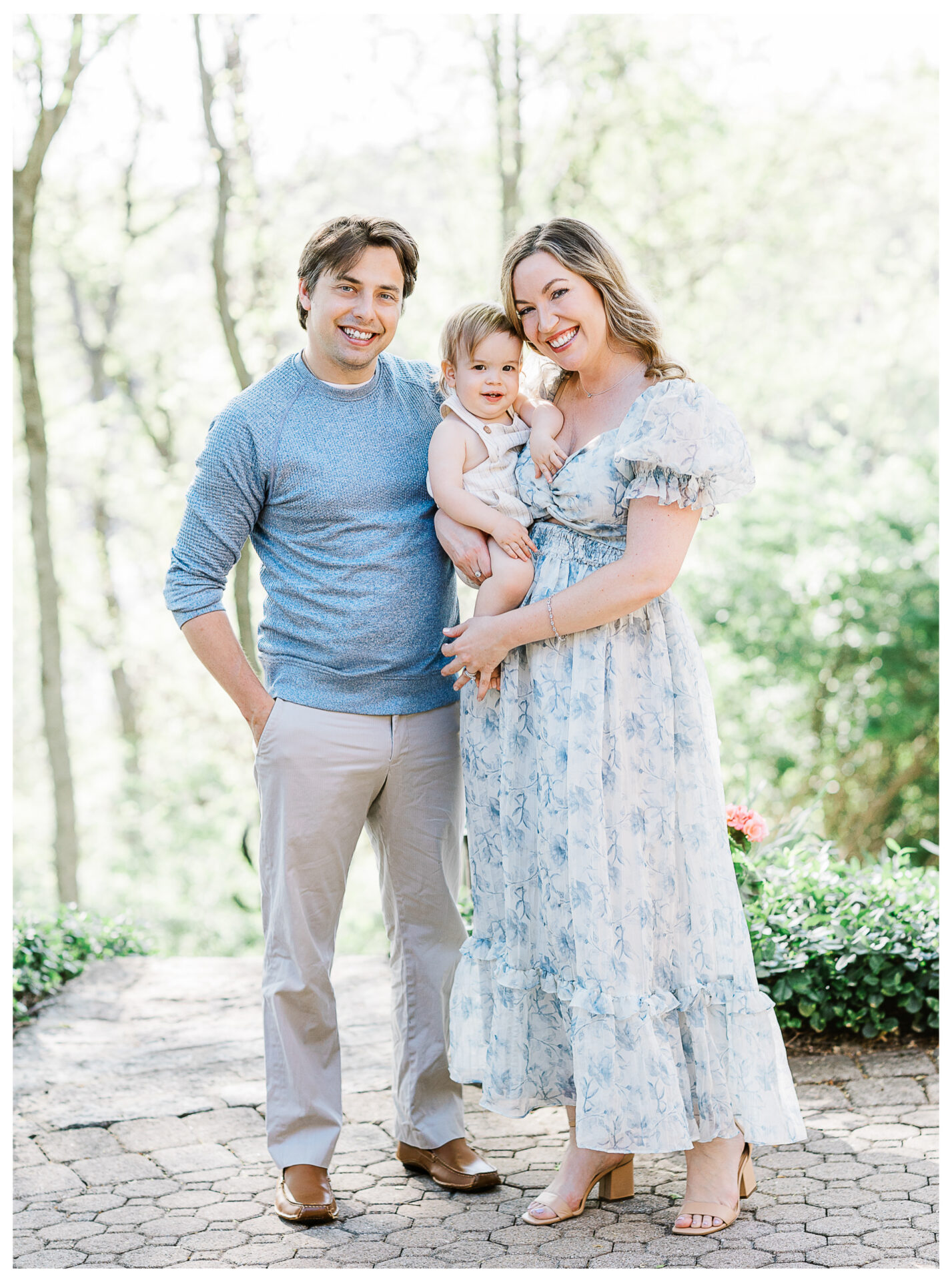 A smiling husband and wife stand outdoors with their baby boy. The man wears a blue sweater and beige pants, the woman wears a light blue floral dress, and the baby is dressed in a light outfit. Trees and greenery are in the background.