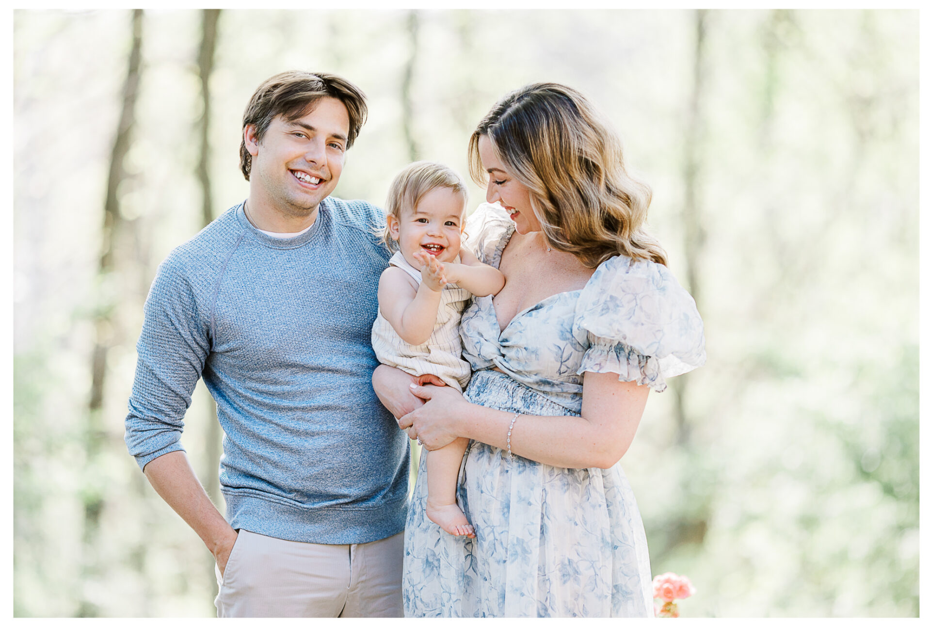 A husband and wife stand outdoors smiling, with the woman holding their happy baby boy dressed in a light outfit. The family is surrounded by soft, natural light and blurred greenery in the background.