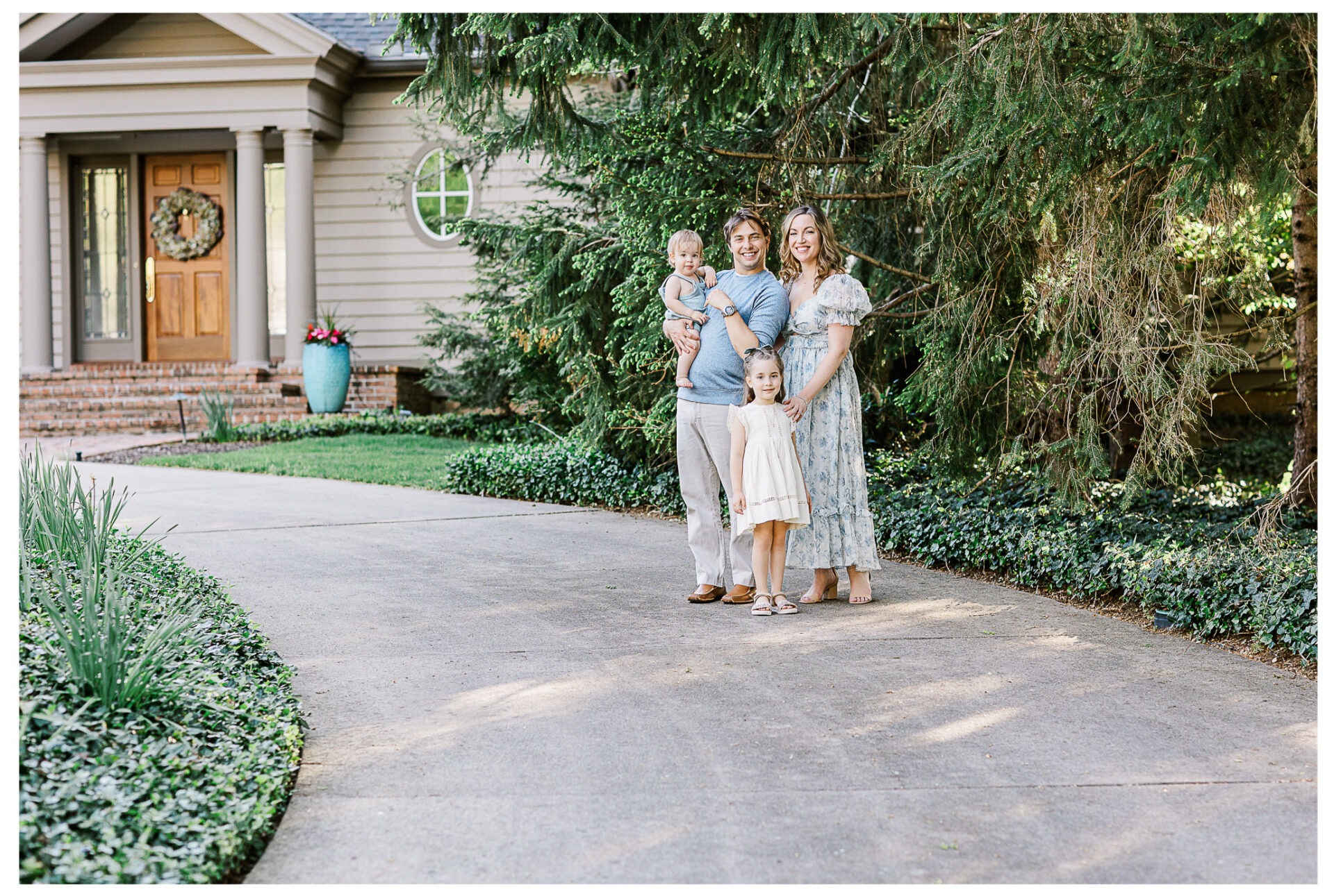 A family of four stands smiling on the driveway in front of their house with a wreath on the door. The father holds his baby son, while the mother stands beside their young daughter in a light dress. Tall trees and greenery surround them.
