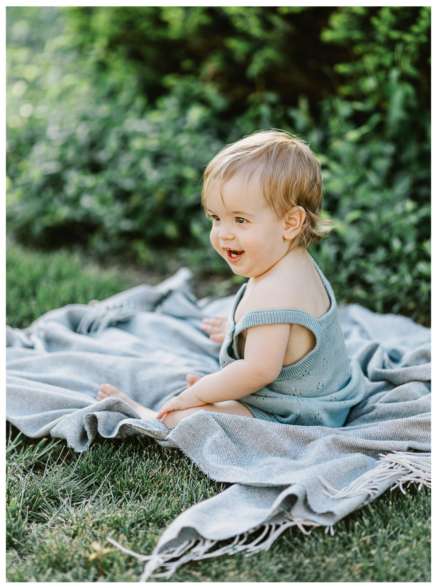 A smiling baby with light hair sits on a gray blanket outdoors on green grass, surrounded by lush greenery, wearing a light blue sleeveless outfit.