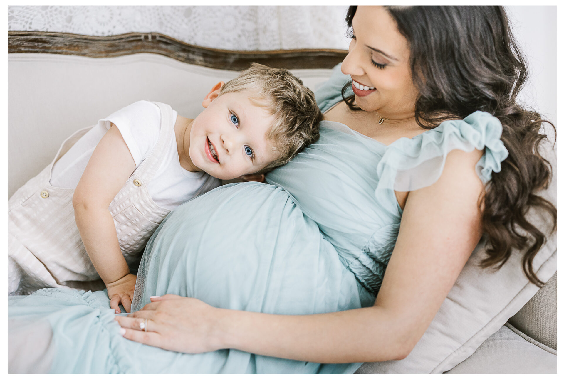 A smiling pregnant mother in a light blue dress sits on a couch as her young son leans his head lovingly on her baby bump, both looking happy and content.
