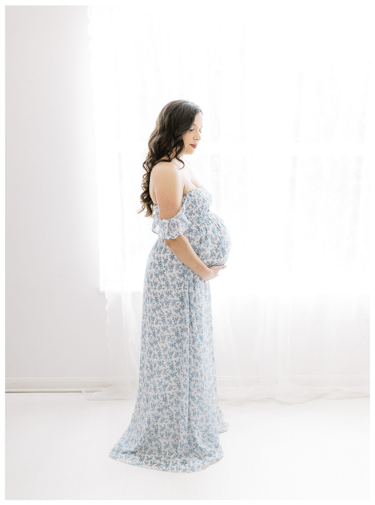 A pregnant woman in a light blue floral dress stands in profile by a bright window with sheer white curtains, gently cradling her belly and looking down softly.