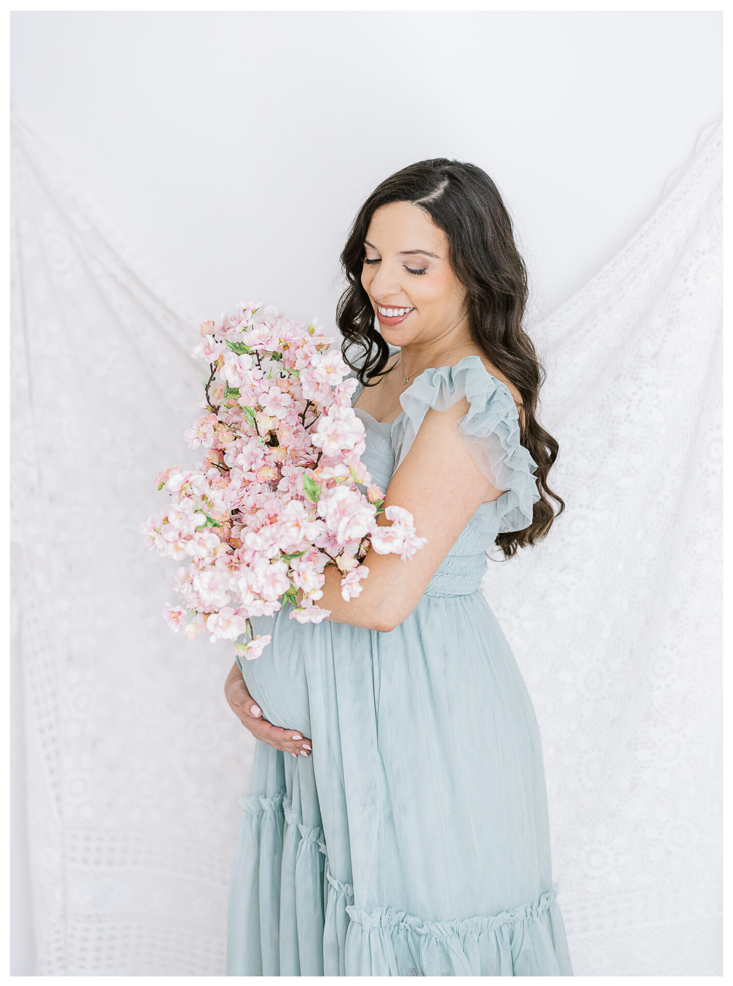 A pregnant woman in a light blue dress holds a bouquet of pink flowers, smiling and looking down, with a white lace fabric as the background.