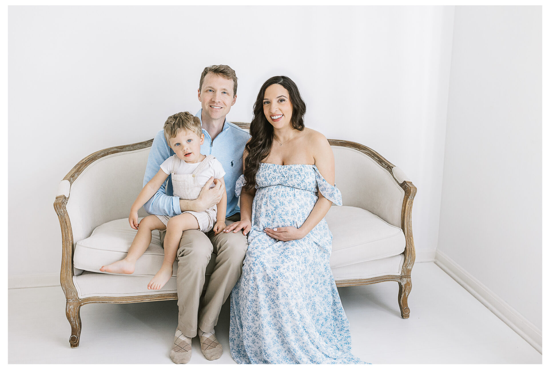 A smiling family of three sits on a cream-colored sofa against a white background. The mother, visibly pregnant and wearing a blue floral dress, sits beside the father, who holds their young son on his lap.