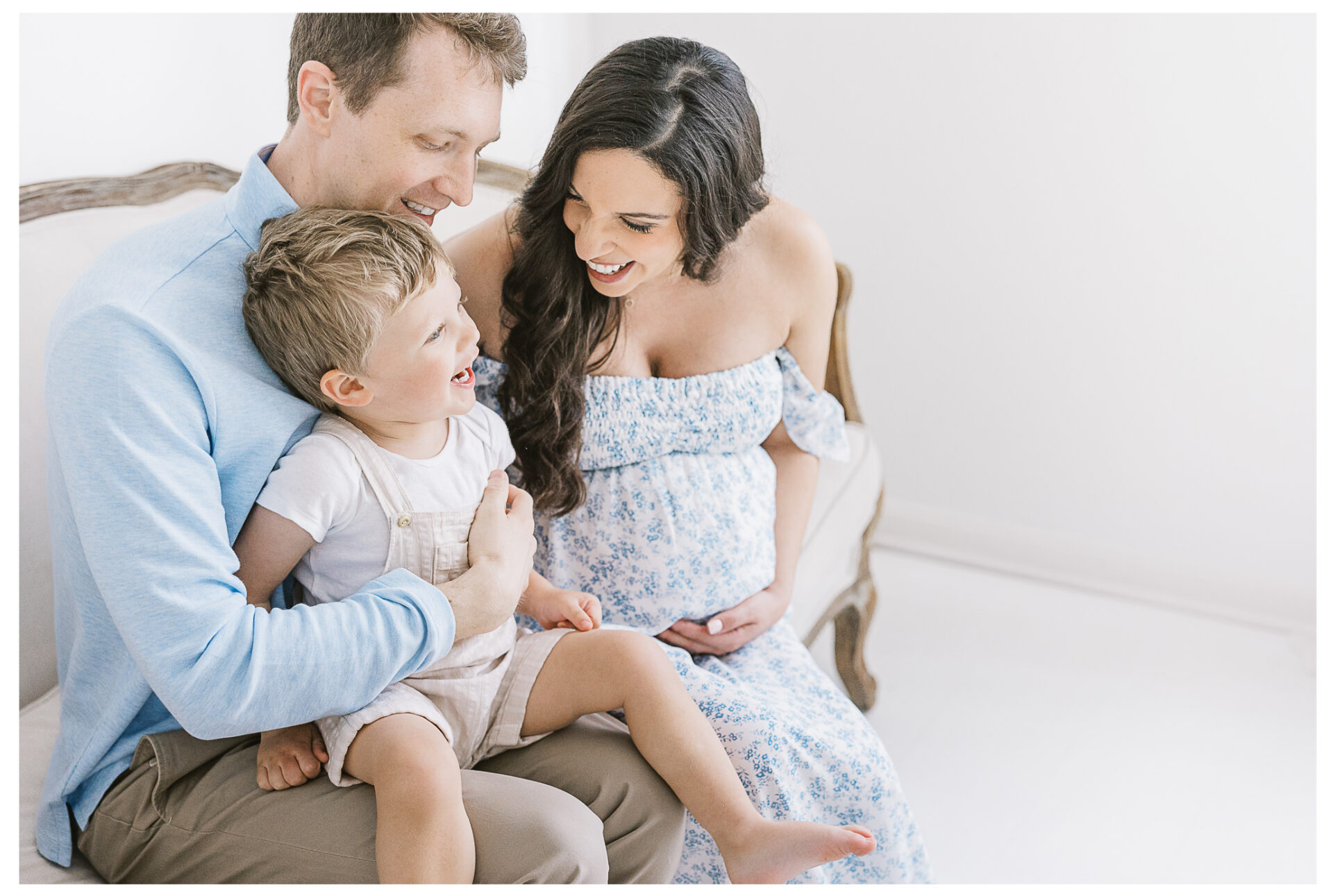 A father and expecting mother sit on a light-colored couch, smiling at each other while holding their young son on their laps. The woman wears a blue floral dress, and the boy looks happy, dressed in light overalls.