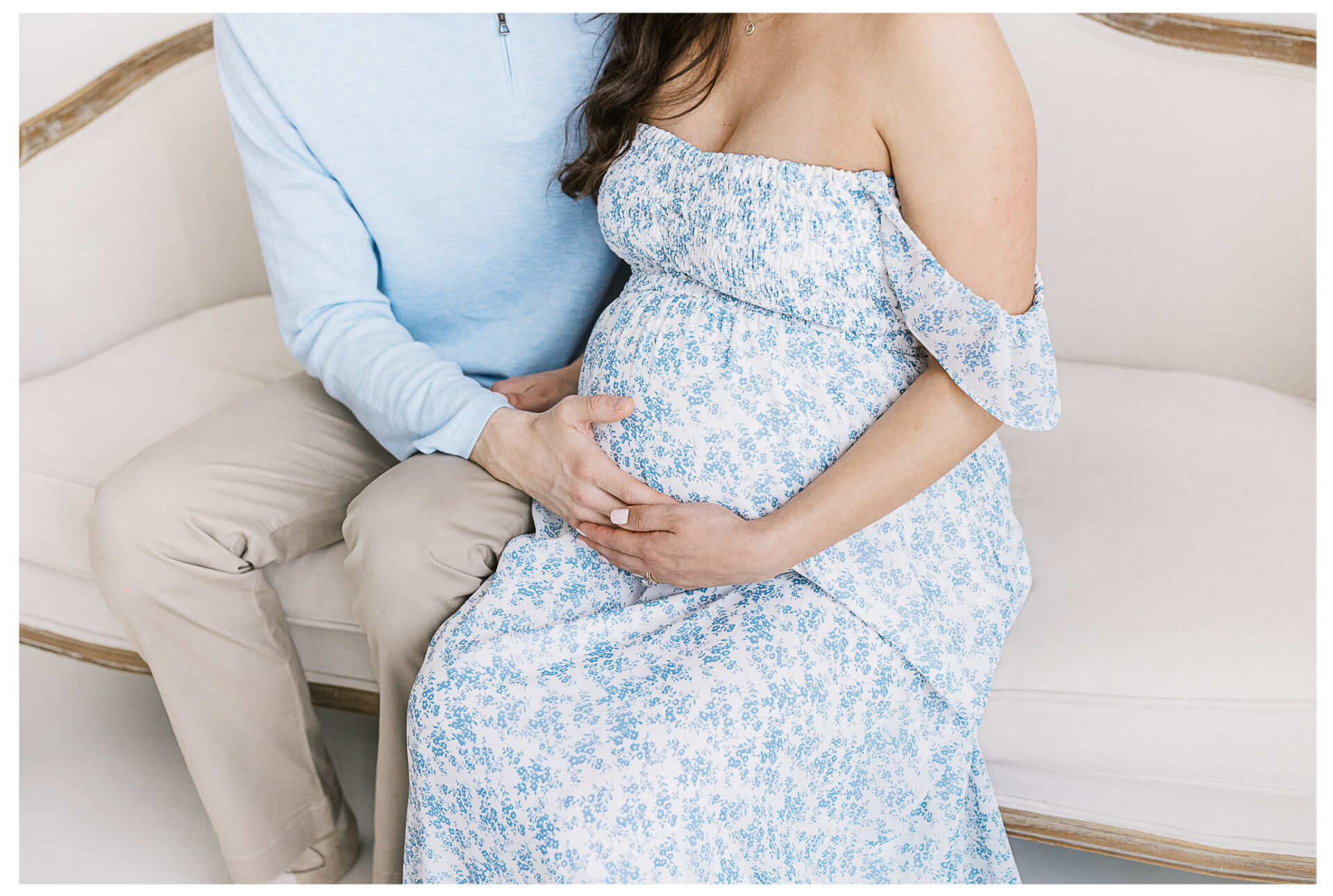 A pregnant woman in a blue floral dress sits on a couch with her husband beside her, wearing light blue and beige, gently rests his hand on her baby bump. Their faces are not visible.